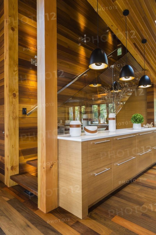 Bamboo wood buffet with white quartz countertop illuminated by black industrial style pendant lighting fixtures next to staircase with clear glass railing in kitchen with Ipe wood floor inside luxurious stained cedar and timber wood home, Quebec, Canada, North America