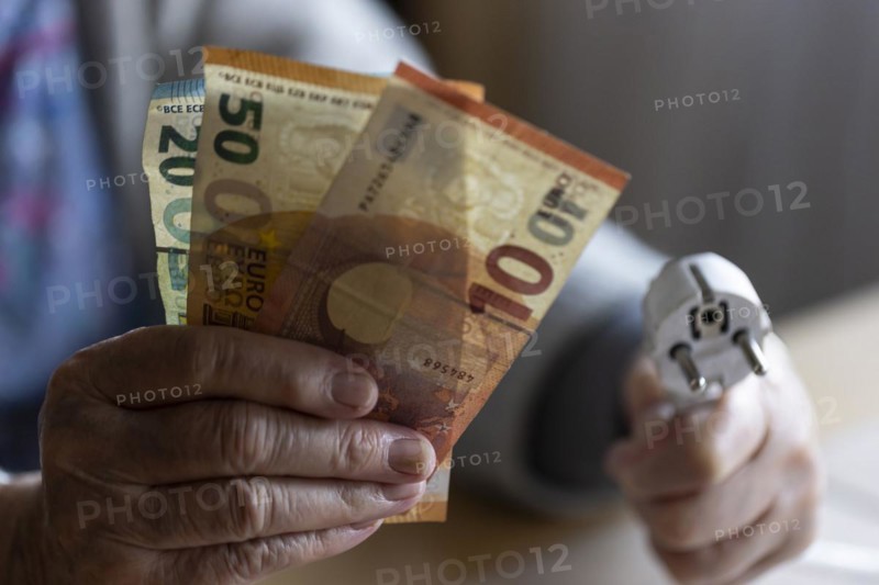 Senior citizen holding money and a power cable with plug in her hand at home, symbolising energy costs and poverty, Cologne, North Rhine-Westphalia, Germany, Europe