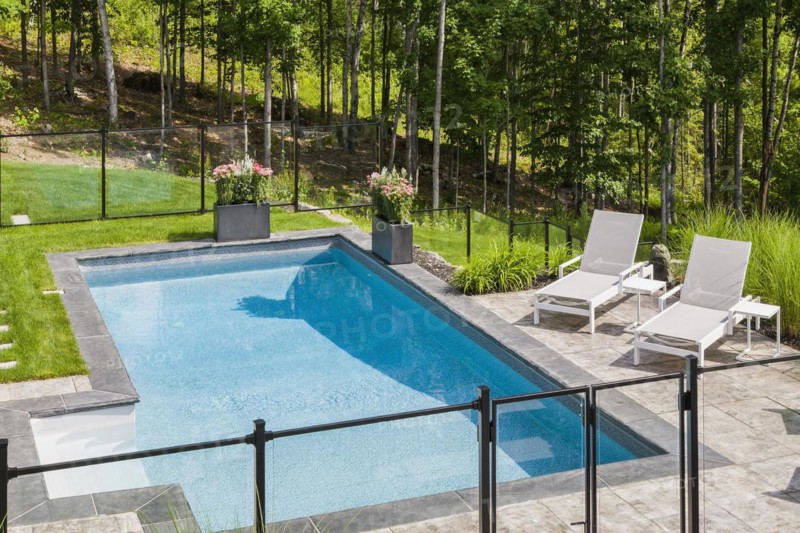 Two white long chairs on edge of in-ground swimming pool enclosed by clear glass and black metal fence in residential backyard in summer, Quebec, Canada, North America