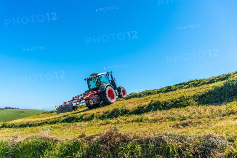 A tractor is driving down a hillside, plowing the grass. The sky is clear and blue, and the sun is shining brightly. The scene is peaceful and serene, with the tractor moving slowly