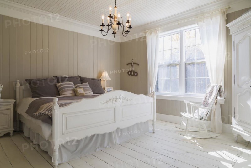 King size bed with light brown and beige striped bedspread, white painted wooden headboard and footboard with carved details, rocking chair and tall armoire in master bedroom with white larch wood floorboards on upstairs floor inside country style home, Quebec, Canada, North America
