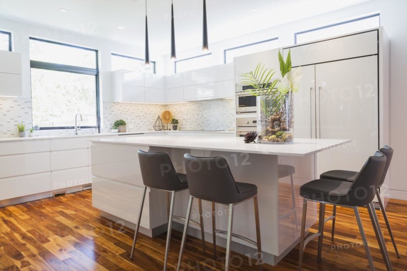 White high-gloss lacquered cabinets and island with grey leather bar stools and white quartz countertops in kitchen with acacia wood floorboards inside contemporary home, Quebec, Canada, North America