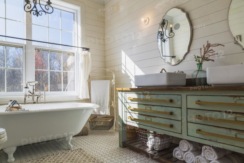 Green antique finish pine wood vanity with rustic wood plank countertop and white top-mounted his and hers vessel sinks, freestanding claw foot bathtub in en suite with honeycomb ceramic tile flooring on upstairs floor inside country style home, Quebec, Canada, North America