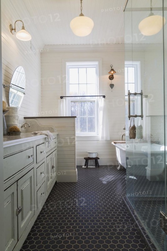 White antique style finish vanity with quartz countertop and two sunken sinks, freestanding claw foot bathtub, glass shower stall in kid's upstairs floor bathroom with black honeycomb ceramic tile floor inside country style home, Quebec, Canada, North America