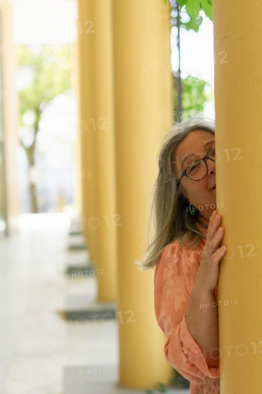 Older woman with white hair and glasses, smiling from behind a column