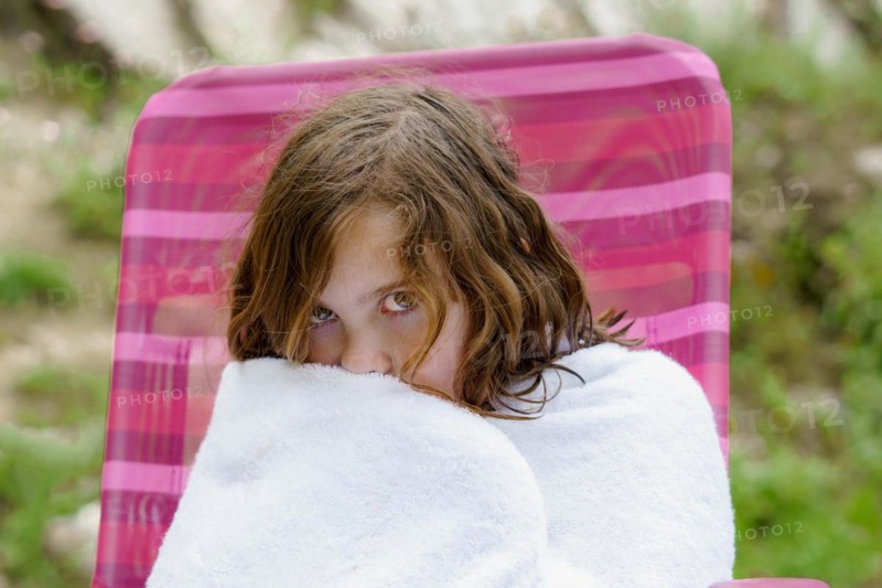 Pretty eight-year-old girl sitting on a chair wrapped in a white towel after getting out of the water, looking cold