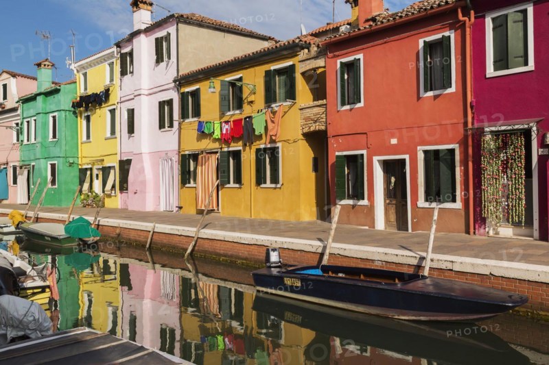 Moored boats on canal lined with pink, red, orange, yellow and green stucco houses decorated with curtains over entrance doors plus washed clothes on clothesline, Burano Island, Venetian Lagoon, Veneto, Italy, Europe