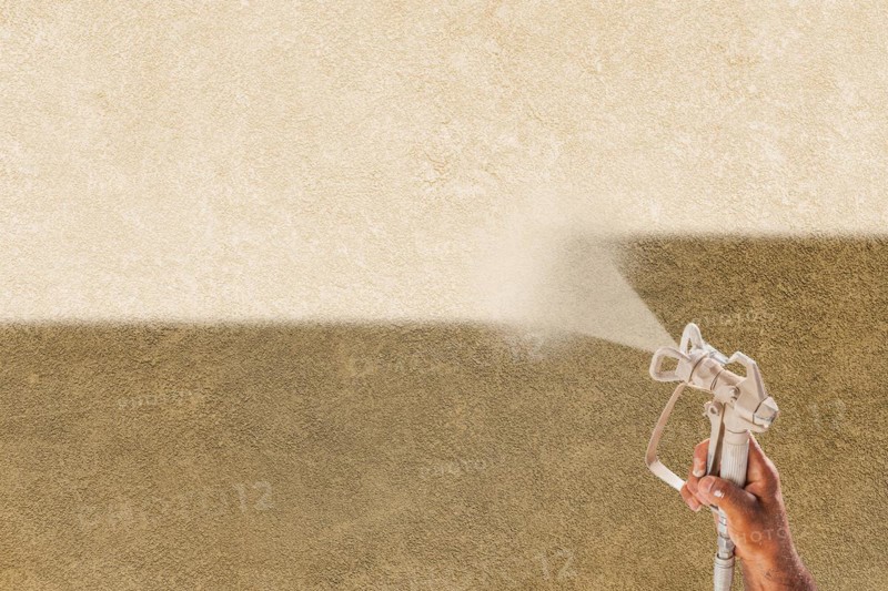 Professional spray painter holding spray gun spraying white paint over brown stucco surface
