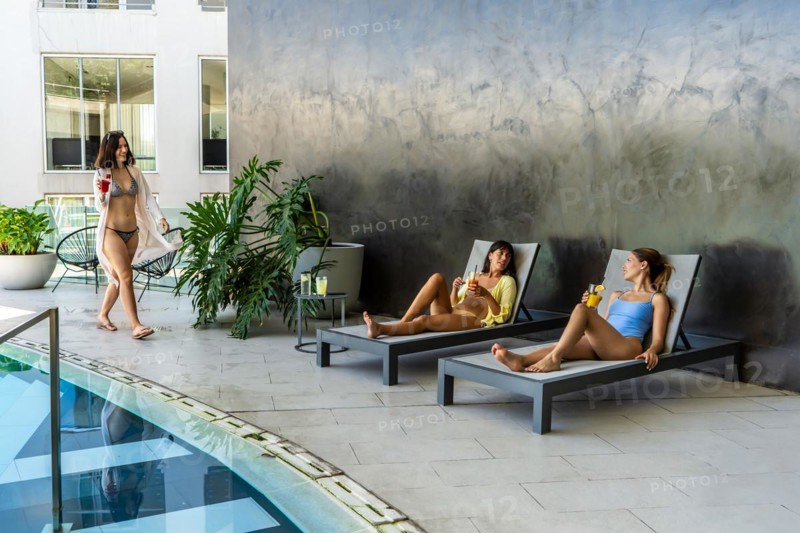 Two female friends are lounging in a fancy hotel pool area, laughing and enjoying themselves while waiting for their other friend to join them