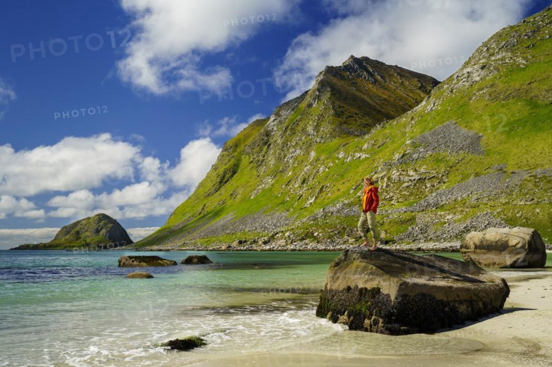 Landscape with sea at the sandy beach of Haukland (Hauklandstranda) with the mountain Veggen. A woman stands barefoot on a rock and looks out to sea. Shot during the day with blue sky. Early summer. Haukland Beach, Haukland, Vestvagoya, Lofoten, Norway, Europe