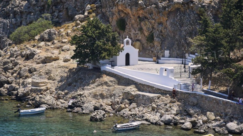 A white church on the Mediterranean Sea on a rocky shore next to parked boats, St Paul's Chapel, Paul's Bay, below the Acropolis of Lindos, Lindos, Rhodes, Dodecanese, Greek Islands, Greece, Europe