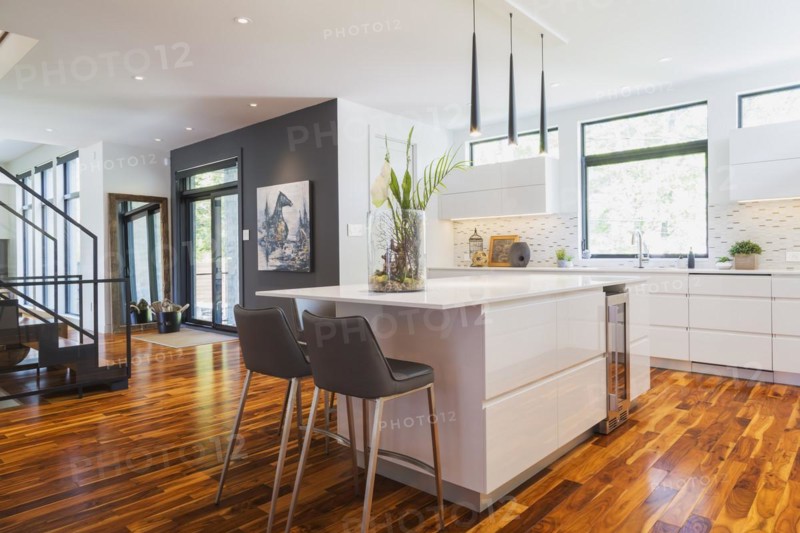 White high-gloss lacquered cabinets and island with grey leather bar stools and white quartz countertops in kitchen with acacia wood floorboards inside contemporary home, Quebec, Canada. Artwork by Liza Castro