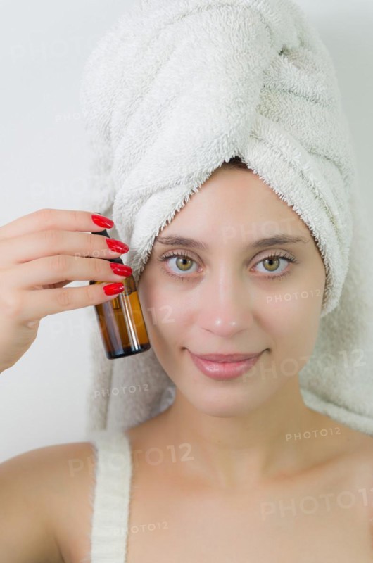 Happy young woman with towel on head holding amber glass bottle of skin serum. Beautiful young Brazilian woman moisturizes her skin with serum isolated against white background