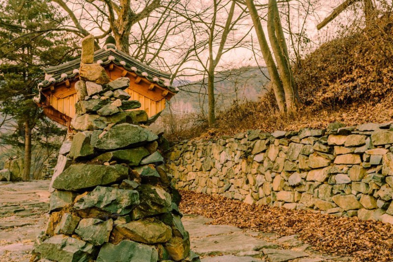 Stone and rock cone shaped tower in front of small wooden oriental building at rural roadside park in South Korea