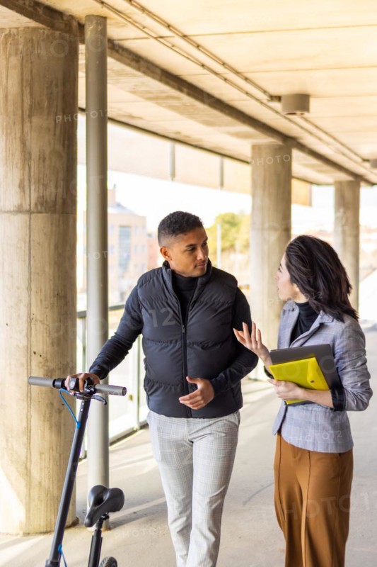 Woman in formal suit talking to man holding a scooter. Girl in formal suit talking to man holding electric scooter while they are walking