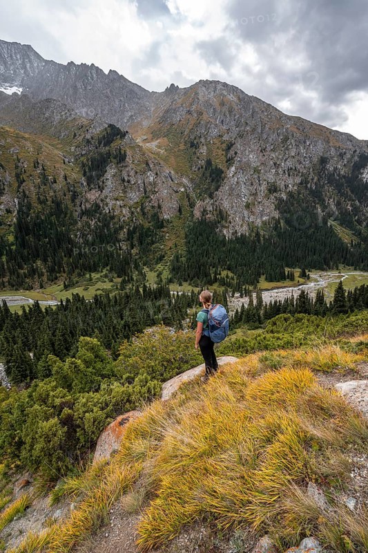 A female hiker stands on a grassy slope against an impressive mountain backdrop under a cloudy sky, Chong Kyzyl Suu Valley, Terskey Ala Too, Tien-Shan Mountains, Kyrgyzstan, Asia