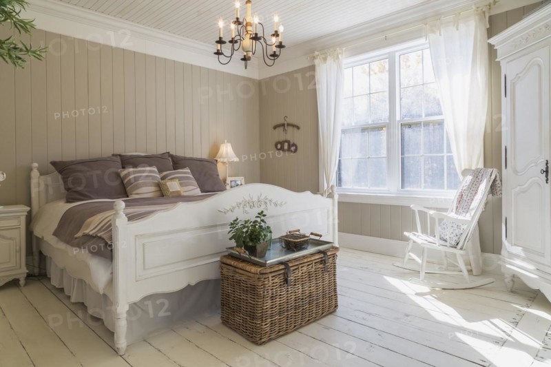 King size bed with light brown and beige striped bedspread, white painted wooden headboard and footboard with carved details, rocking chair and tall armoire in master bedroom with white larch wood floorboards on upstairs floor inside country style home, Quebec, Canada, North America