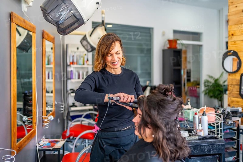 Smiling female hairdresser flat ironing hair of a woman sitting in front of a mirror in the salon