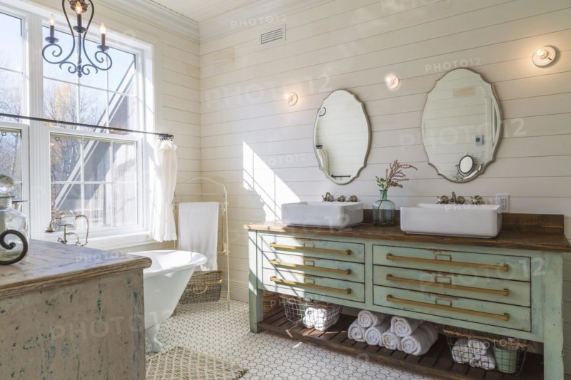 Green antique finish pine wood vanity with rustic wood plank countertop and white top-mounted his and hers vessel sinks, freestanding claw foot bathtub, distressed cabinet in en suite with honeycomb ceramic tile flooring on upstairs floor inside country style home, Quebec, Canada, North America