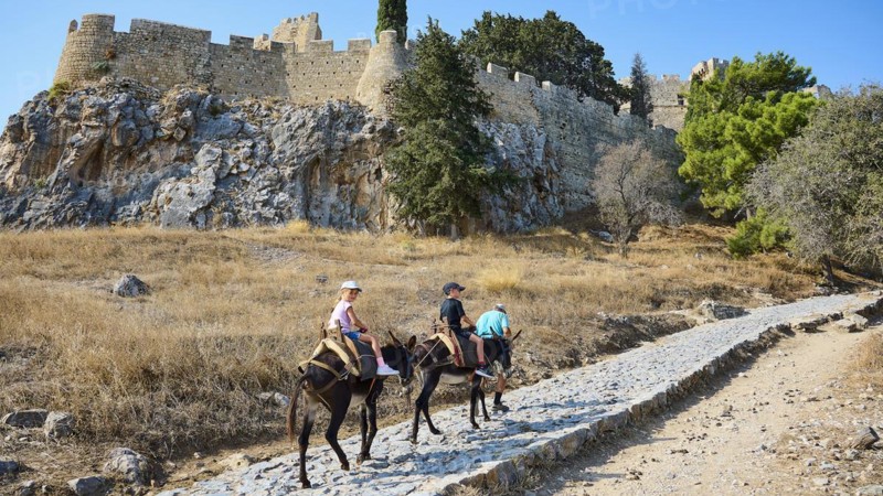 Donkey trail to the Acropolis, children riding donkeys on a dusty hiking trail in front of an old fortress, Lindos, Rhodes, Dodecanese, Greek Islands, Greece, Europe