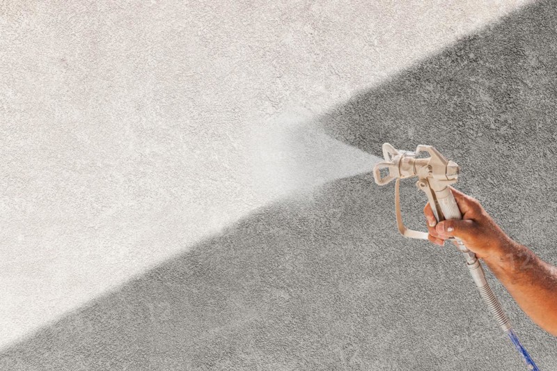Professional spray painter holding spray gun spraying white paint over grey stucco surface