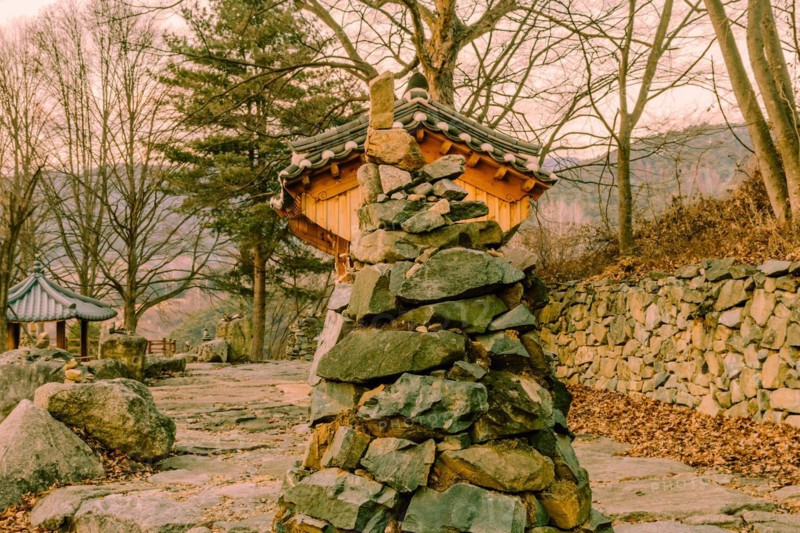 Stone and rock cone shaped tower in front of small wooden oriental building at rural roadside park in South Korea