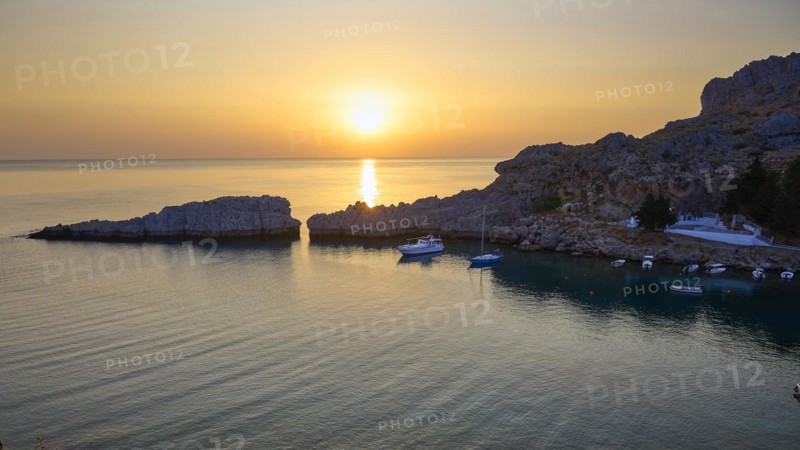 Golden glowing sunset over the calm sea next to rocky cliffs, Paulus Bay, below the Acropolis of Lindos, Lindos, Rhodes, Dodecanese, Greek Islands, Greece, Europe