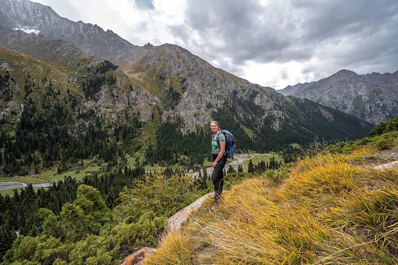 A female hiker stands on a grassy slope against an impressive mountain backdrop under a cloudy sky, Chong Kyzyl Suu Valley, Terskey Ala Too, Tien-Shan Mountains, Kyrgyzstan, Asia