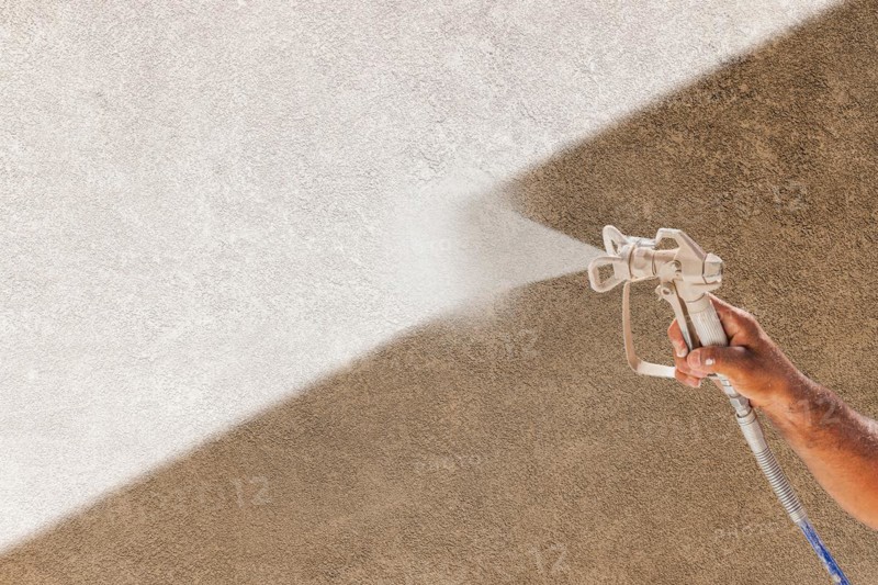 Professional spray painter holding spray gun spraying white paint over brown stucco surface