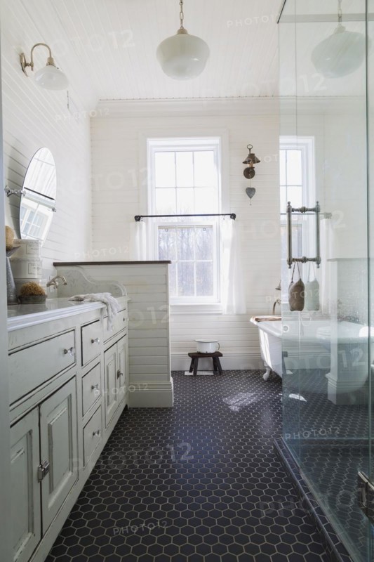 White antique style finish vanity with quartz countertop and two sunken sinks, freestanding claw foot bathtub, glass shower stall in kid's upstairs floor bathroom with black honeycomb ceramic tile floor inside country style home, Quebec, Canada, North America