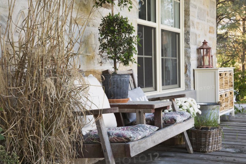 Weathered wood sitting bench with cushions plus various plants, herbs including ornamental grass plant and decorations on front veranda of country style home in autumn, Quebec, Canada, North America