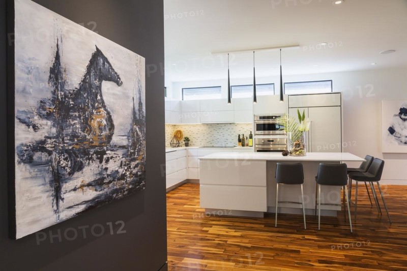Decorative acrylic painting of mystic horse and castle by Liza Castro-Interior Designer on dark living room wall plus white high-gloss lacquered cabinets and island with grey leather bar stools and white quartz countertops in kitchen with acacia wood floorboards inside contemporary home, Quebec, Canada. Artwork by Liza Castro