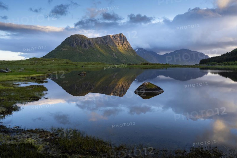 Landscape on the Lofoten Islands. The mountain Offersoykammen and the Beech Vagspollen. Mount Stornappstinden on Flakstadoya in the background. The mountains are reflected in the water. At night during the midnight sun. A few clouds in the sky. The night sun shines on the mountain peak. Early summer. Vestvagoya, Lofoten, Norway, Europe