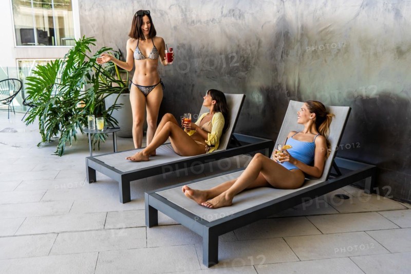 Two women are lounging in a courtyard, while the third is standing beside them. They are sipping on juices while chatting