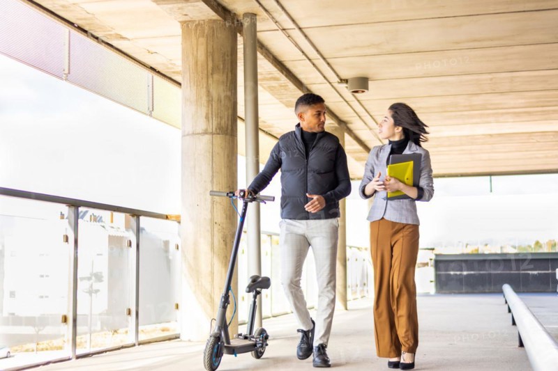 Woman in formal suit talking to man holding a scooter. Girl in formal suit talking to man holding electric scooter while they are walking
