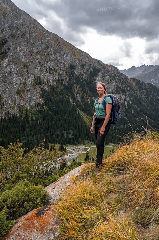 A female hiker stands on a grassy slope against an impressive mountain backdrop under a cloudy sky, Chong Kyzyl Suu Valley, Terskey Ala Too, Tien-Shan Mountains, Kyrgyzstan, Asia