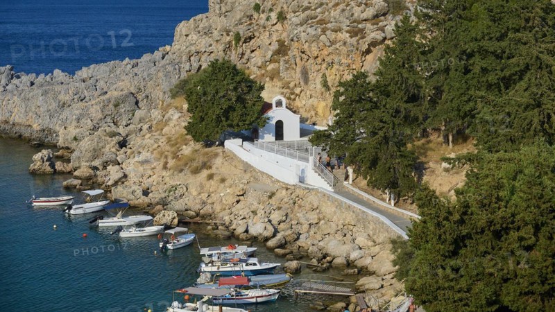 Small church on a rocky path surrounded by boats on a clear sea, St Paul's Chapel, Paul's Bay, below the Acropolis of Lindos, Lindos, Rhodes, Dodecanese, Greek Islands, Greece, Europe