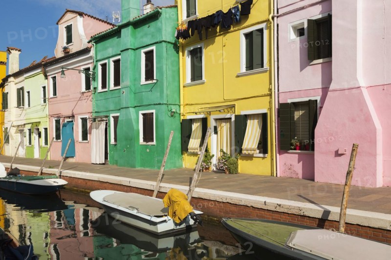 Moored boats on canal lined with pink, yellow and green stucco houses decorated with striped curtains over entrance doors and windows plus clothes on clothesline, Burano Island, Venetian Lagoon, Venice, Veneto, Italy, Europe