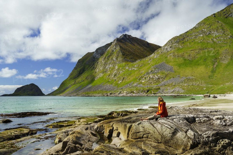 Landscape with sea at the sandy beach of Haukland (Hauklandstranda) with the mountain Veggen. A woman sits barefoot on a rock and looks out to sea. Shot during the day with blue sky. Early summer. Haukland Beach, Haukland, Vestvagoya, Lofoten, Norway, Europe