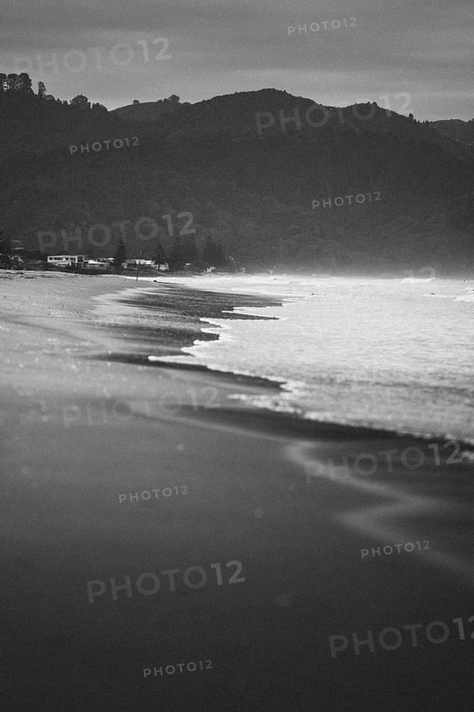 Walk on the beach at sunset, restless sea lapping waves on the beach, rocky landscape in the background. Taken in black and white at Waihi Beach in New Zealand