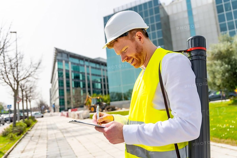 Smart architect writing notes standing in the city wearing safety yellow reflective waistcoat, helmet and holding a draw tube