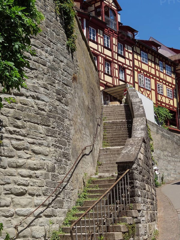 A steep staircase leads next to a high stone wall to a half-timbered house in a sunny old town, steep staircase in a historic town with half-timbered houses, meersburg, germany
