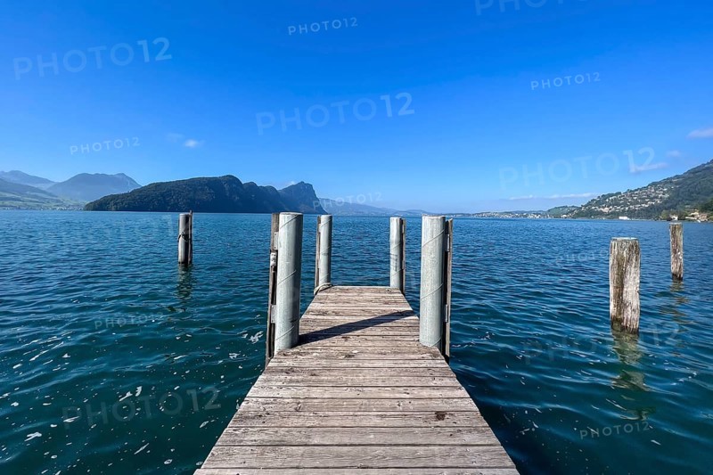 Wooden jetty juts into clear lake under blue sky, surrounded by mountains, Vitznau, Lucerne, Switzerland, Europe