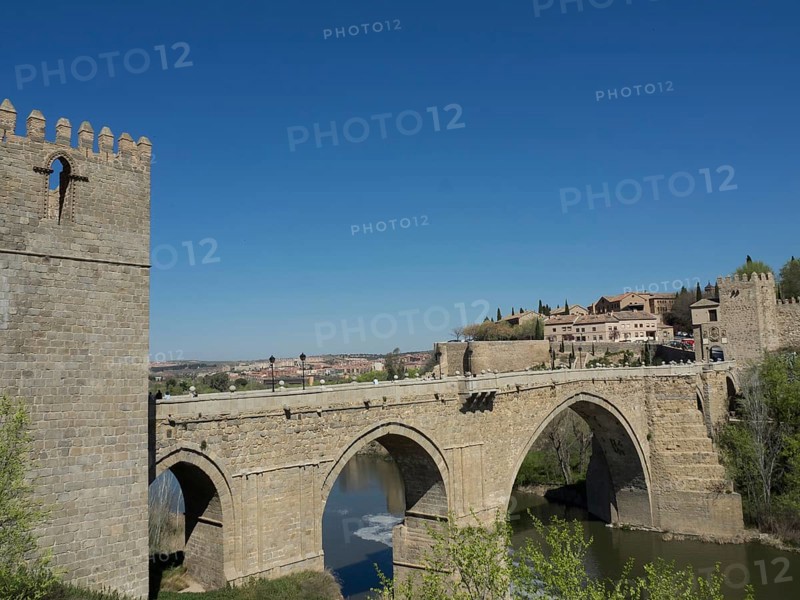 Historic stone bridge over a river with city walls and buildings in the background under a sunny blue sky, historic stone bridge with an old tower and an old city in the background over a wild river, Toledo, Spain, Europe