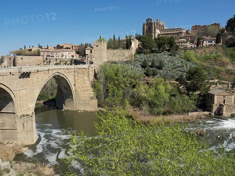 Stone bridge over the river, green hills with historical buildings and cathedral, clear blue sky, historical stone bridge with an old tower and an old city in the background over a wild river, Toledo, Spain, Europe