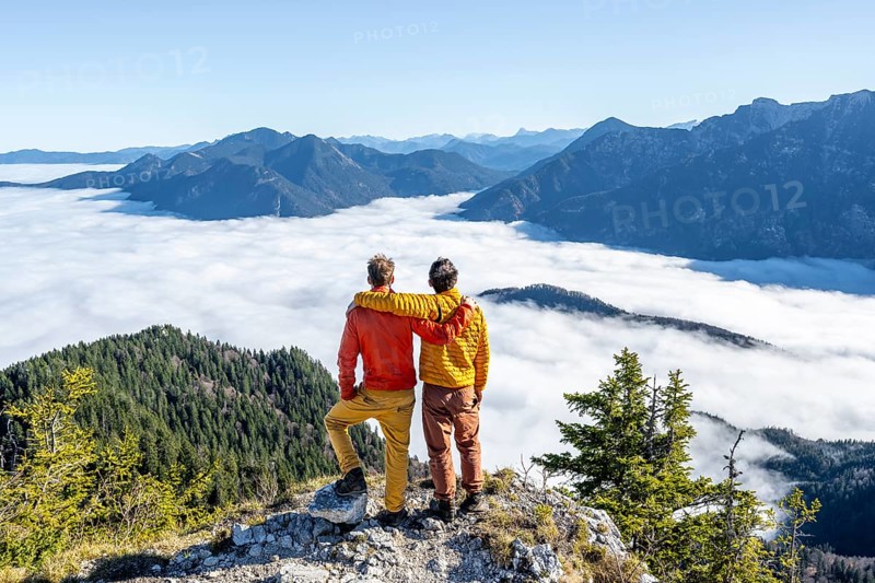 Two friends put their arms around each other's shoulders, mountaineers on the rocky summit of the Ettaler Manndl, view over mountain landscape and sea of clouds, high fog in the valley, Ammergau Alps, Bavaria, Germany, Europe