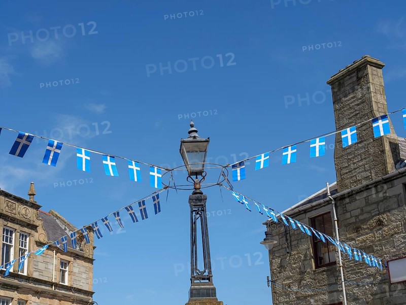 Street scene with blue and white flags hanging from a lamppost between old buildings, calm sea with rocks and fields with historic houses, Lerwick, Shetlands, Scotland, United Kingdom, Europe
