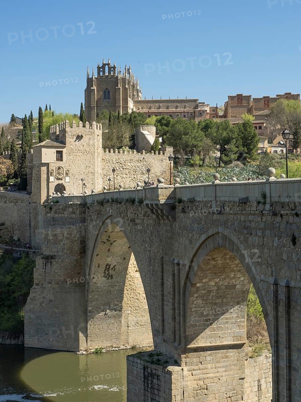 Stone bridge with arches over a river, in the background a church and cityscape, sunny weather, historic stone bridge with an old tower and an old town in the background over a wild river, Toledo, Spain, Europe