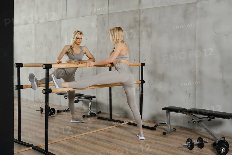 Fitness athlete standing in front of a wall mirror and stretching leg using handrail