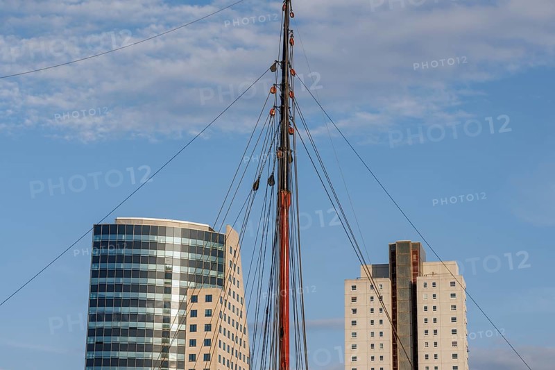 Sail mast between two modern buildings in front of a blue sky, skyline of a modern city on a river with a modern bridge, Rotterdam, Netherlands